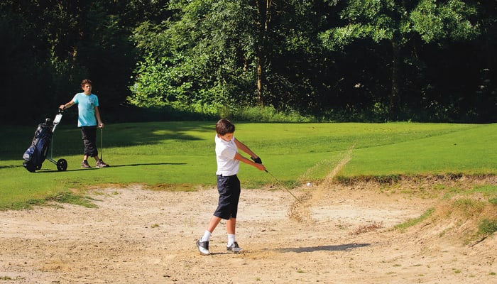 Jeune golfeur en swing sur un parcours de golf avec caddie et sac de golf, entouré de verdure et bunker de sable