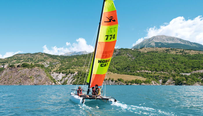 Catamaran de kitesurf coloré aux voiles rouges et jaunes naviguant sur un lac alpin aux Crots avec montagnes en arrière-plan