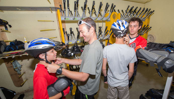 Moniteur VTT descente avec jeunes ados en atelier d'équipement aux Deux Alpes, casques et vélos en arrière-plan