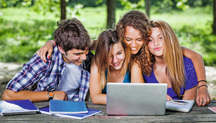 Groupe de quatre adolescents souriants travaillant ensemble sur un ordinateur portable en plein air à Bois-le-Roi