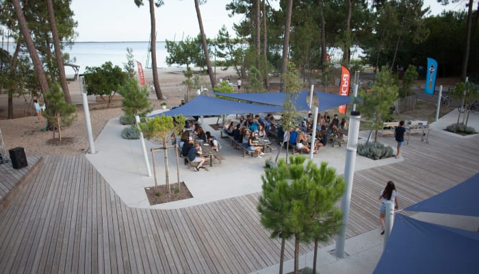 Espace de convivialité à Bombannes avec terrasse ombragée, lac en arrière-plan et visiteurs détendus sous voiles bleues