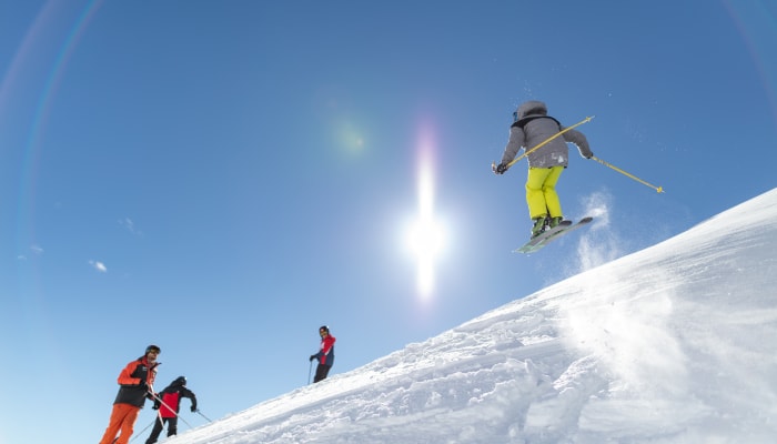 Skieur en pantalon jaune fluorescent effectuant un saut en hors-piste avec groupe d'instructeurs en pente enneigée sous ciel bleu