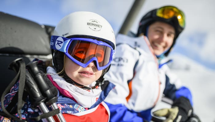 Enfant débutant en ski à la Plagne portant casque blanc et masque bleu avec moniteur souriant en arrière-plan