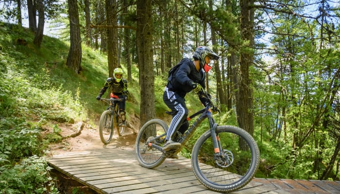Deux vététistes en descente sur un sentier forestier équipé de structures en bois, portant équipement de protection