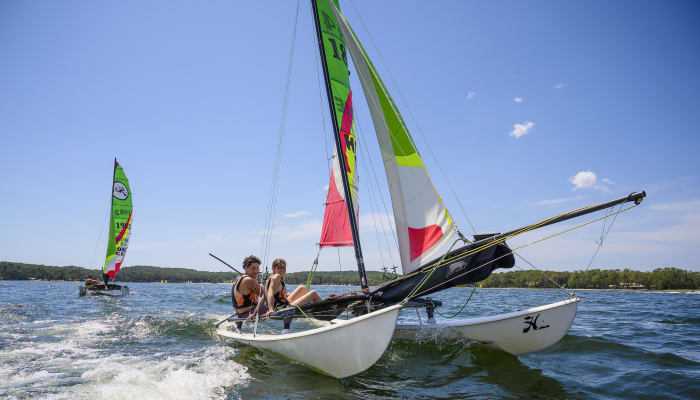 Adolescents naviguant en catamaran lors d'un séjour multisports nautique à Bombannes avec voiles colorées et forêt en arrière-plan