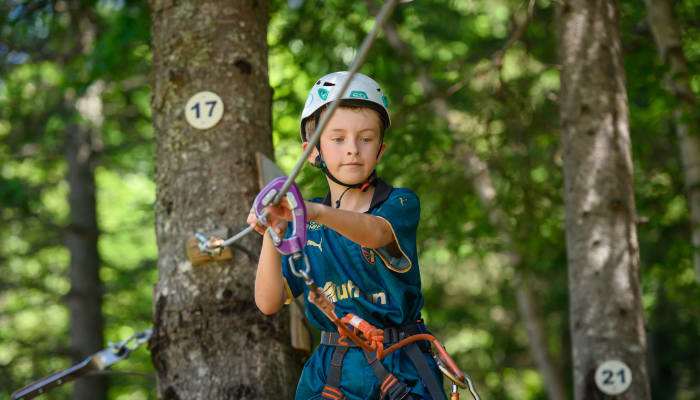 Enfant équipé de casque et harnais pratiquant l'escalade en forêt sur parcours d'aventure multisports