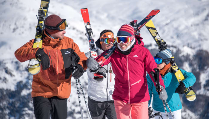 Groupe de quatre skieurs et snowboarders souriants à Serre Chevalier avec skis et snowboards levés, paysage montagneux enneigé en arrière-plan