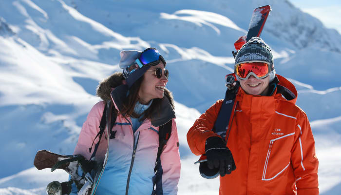 Deux skieurs souriants en tenue colorée dans un paysage montagneux enneigé, équipés de skis et de casques de protection