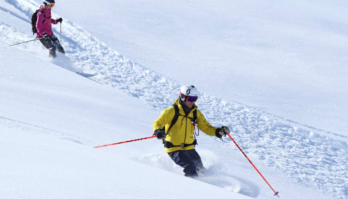 Skieur expert en veste jaune descendant une pente hors-piste enneigée avec moniteur en arrière-plan dans le Briançonnais