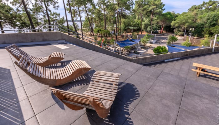 Terrasse avec chaises longues en bois face aux terrains de sport et lac de Bombannes entouré de pins