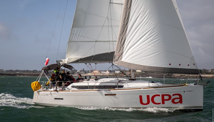 Voilier blanc UCPA naviguant en Morbihan avec équipage à bord et voiles gonflées, archipel côtier en arrière-plan