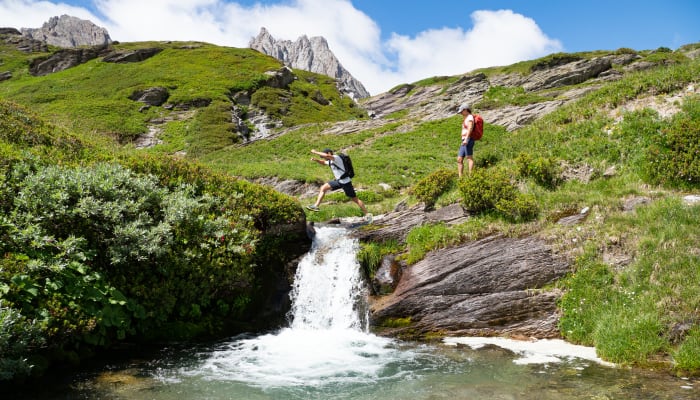 Randonneurs traversant un ruisseau alpin dans les prairies vertes de Serre Chevalier avec pics rocheux en arrière-plan