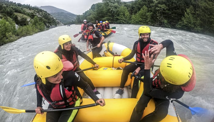 Groupe de jeunes en rafting sur le lac de Serre-Ponçon avec casques jaunes et gilets de sauvetage rouges, entouré de montagnes verdoyantes