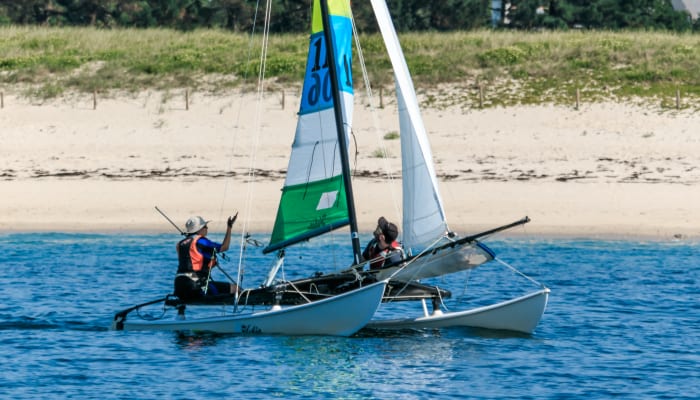 Catamaran à voile avec deux participants en mer, voile multicolore jaune et verte, plage et forêt en arrière-plan