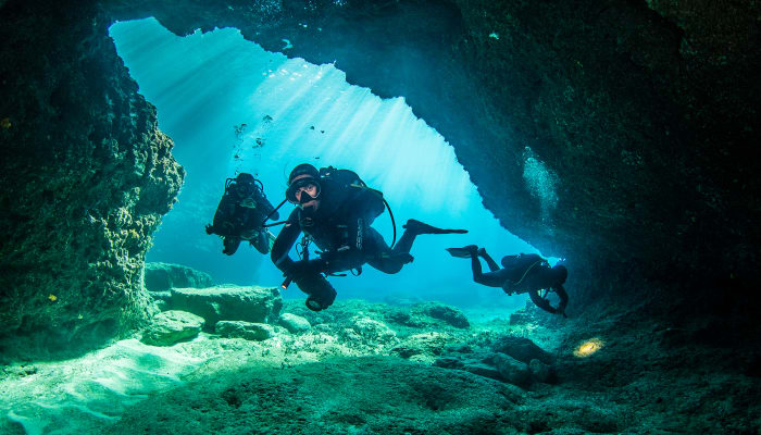 Plongeurs équipés de bouteilles explorant une grotte sous-marine avec rayons de lumière bleue traversant l'eau cristalline