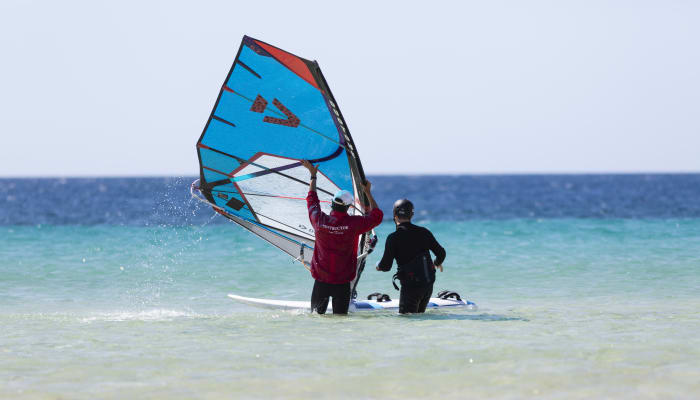 Deux pratiquants de windsurf en mer avec voile bleue et rouge, préparant leur sortie en eau turquoise et peu profonde