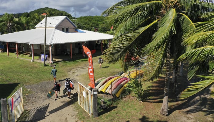 Base UCPA de windsurf avec planches colorées, palmiers et moniteurs accueillant les stagiaires sous le soleil tropical