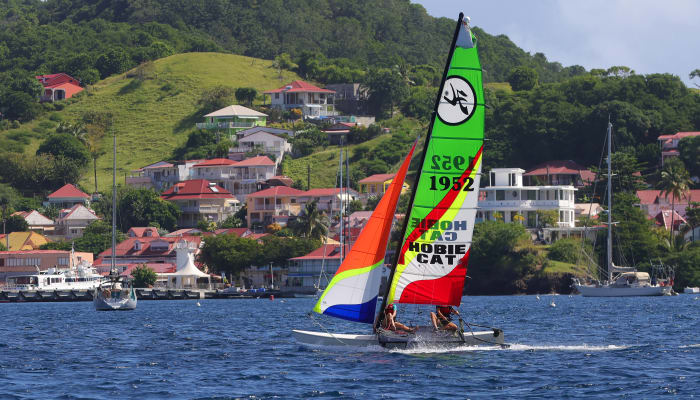 Catamaran aux voiles colorées naviguant dans la baie des Saintes avec îles verdoyantes et village côtier en arrière-plan