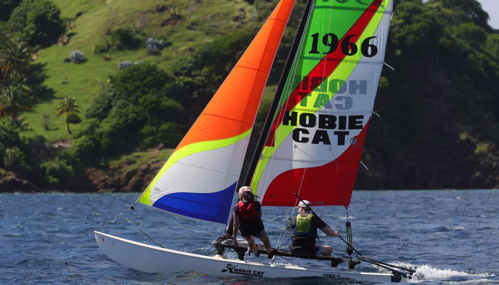 Catamaran avec voiles colorées naviguant en baie des Saintes, deux navigateurs à bord, paysage tropical verdoyant en arrière-plan