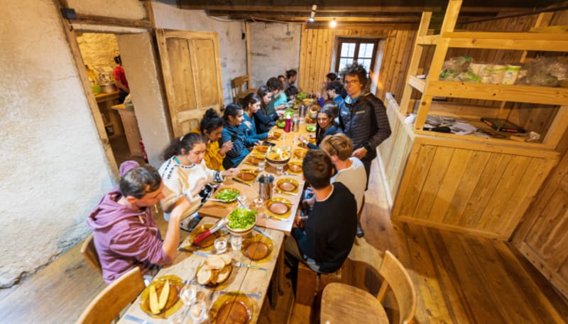 Jeunes randonneurs partageant un repas en refuge montagnard avec table en bois et lits superposés