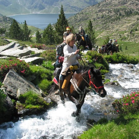 Randonnée équestre en montagne traversant un ruisseau avec lac et vallée pyrénéenne en arrière-plan