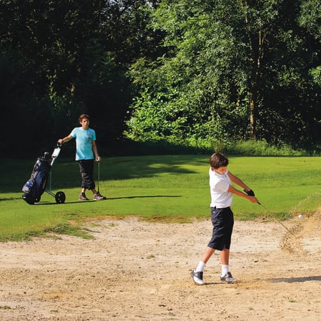 Jeune golfeur en swing sur un parcours de golf avec caddie et sac de golf, entouré de verdure et bunker de sable
