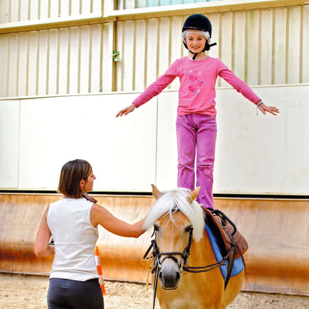 Jeune cavalière en équilibre sur un poney blanc, portant casque et tenue rose, dans une carrière équestre couverte