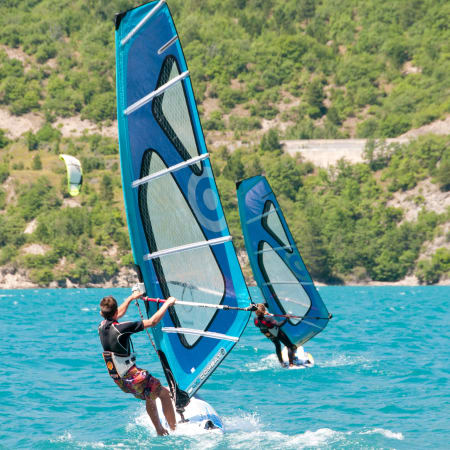 Pratiquants de wingsurf en action sur eau turquoise avec voiles bleues et paysage montagneux boisé en arrière-plan