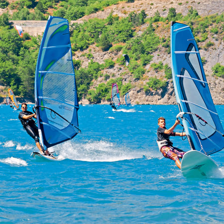 Jeunes windsurfeurs pratiquant le windsurf entre rivière et lac avec voiles bleues et falaises rocheuses en arrière-plan