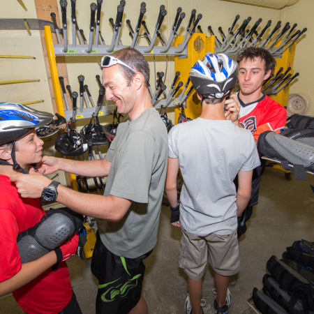 Moniteur VTT descente avec jeunes ados en atelier d'équipement aux Deux Alpes, casques et vélos en arrière-plan