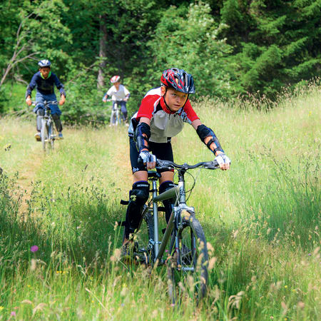 Cycliste en VTT au premier plan sur sentier herbeux avec groupe de riders en montagne boisée