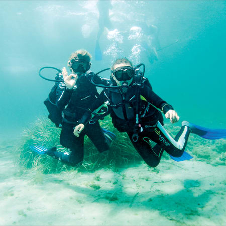 Deux plongeurs en combinaison et équipement complet explorant le fond marin avec coraux et faune sous-marine