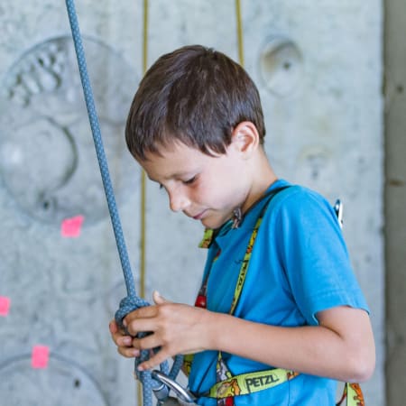 Enfant en escalade intérieure tenant la corde avec harnais et équipement de sécurité sur mur gris