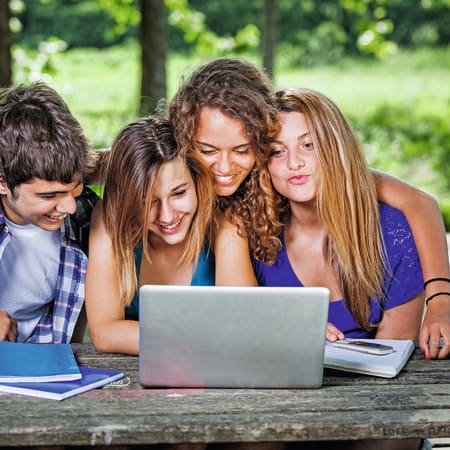 Groupe de quatre adolescents souriants travaillant ensemble sur un ordinateur portable en plein air à Bois-le-Roi