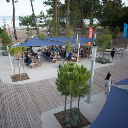 Espace de convivialité à Bombannes avec terrasse ombragée, lac en arrière-plan et visiteurs détendus sous voiles bleues