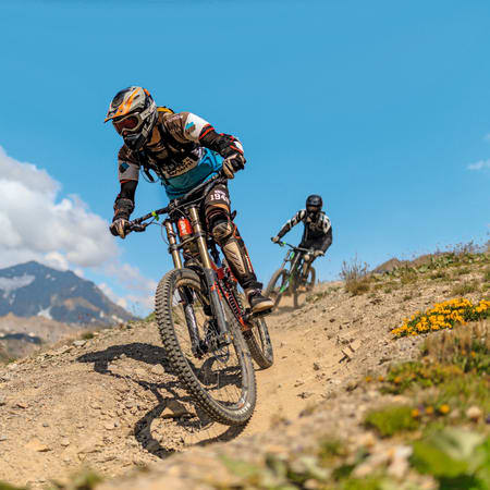 Cycliste en descente sur VTT en montagne avec équipement de protection, paysage alpin et autre rider en arrière-plan