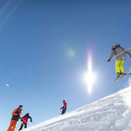 Skieur en pantalon jaune fluorescent effectuant un saut en hors-piste avec groupe d'instructeurs en pente enneigée sous ciel bleu