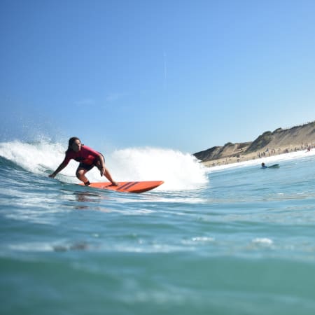 Surfeur en combinaison rouge équilibré sur sa planche orange chevauchant une vague, plage de sable fin en arrière-plan