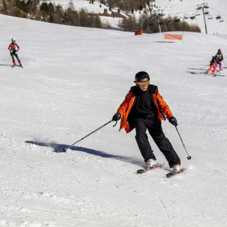 Jeune skieur en veste orange dévale les pentes enneigées des Orres avec d'autres skieurs et snowboarders en arrière-plan