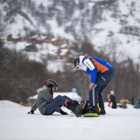 Moniteur en tenue bleue et orange aidant un jeune snowboarder au sol à Saint-Lary-Soulan avec montagnes enneigées en arrière-plan