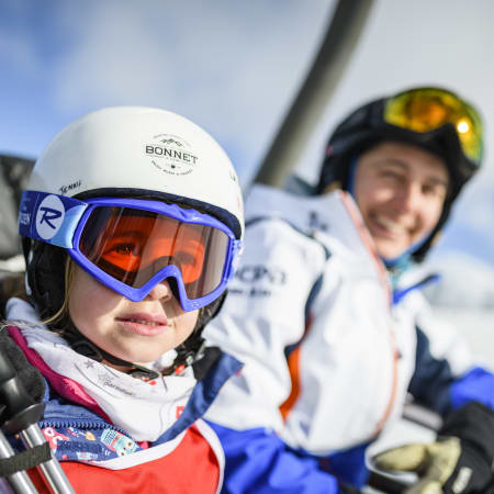 Enfant débutant en ski à la Plagne portant casque blanc et masque bleu avec moniteur souriant en arrière-plan