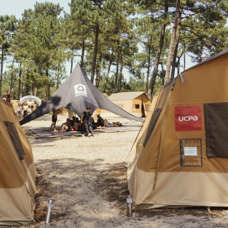 Campement de surf avec tentes bêches et zone commune sous auvent noir entre pins, accueil pour jeunes surfeurs