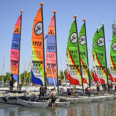 Flotte de catamarans colorés avec voiles orange, rose, bleu et vert amarrés à proximité d'une école de voile, prêts pour l'entraînement