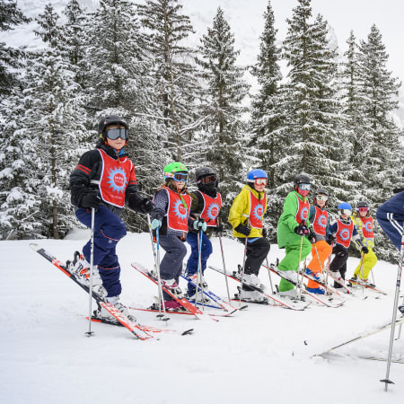 Groupe d'enfants en tenue de ski colorée suivant un moniteur sur piste enneigée avec sapins en arrière-plan