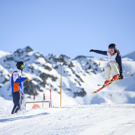 Jeune skieur en vol effectuant une figure freestyle aux Arcs avec moniteur observant, pistes enneigées et montagnes en arrière-plan