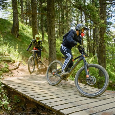 Deux vététistes en descente sur un sentier forestier équipé de structures en bois, portant équipement de protection