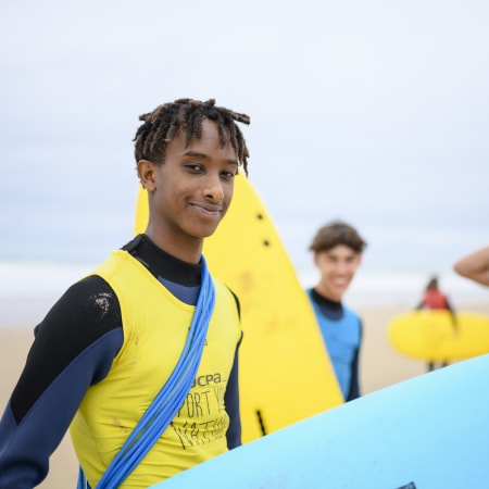 Adolescent souriant tenant sa planche de surf jaune lors d'un séjour surf à Bombannes avec d'autres jeunes en arrière-plan