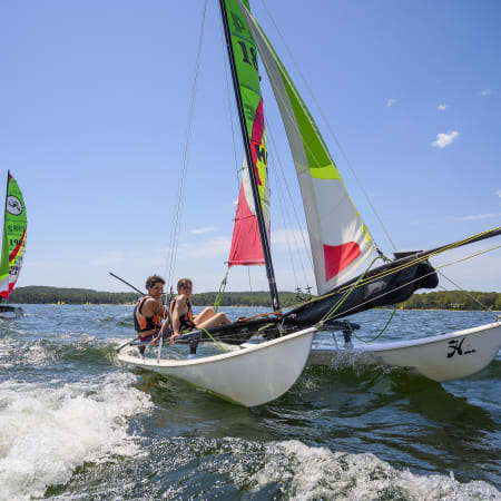 Adolescents naviguant en catamaran lors d'un séjour multisports nautique à Bombannes avec voiles colorées et forêt en arrière-plan