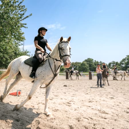 Enfant en équitation galopant sur un cheval blanc en carrière avec d'autres cavaliers en arrière-plan lors d'un stage multisports à Valloire