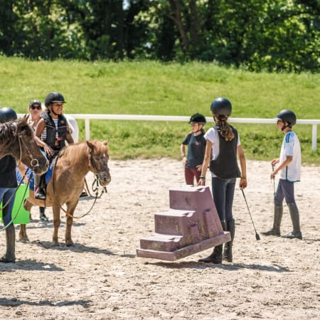 Moniteur UCPA guidant des jeunes cavaliers lors d'une leçon d'équitation en carrière avec obstacles